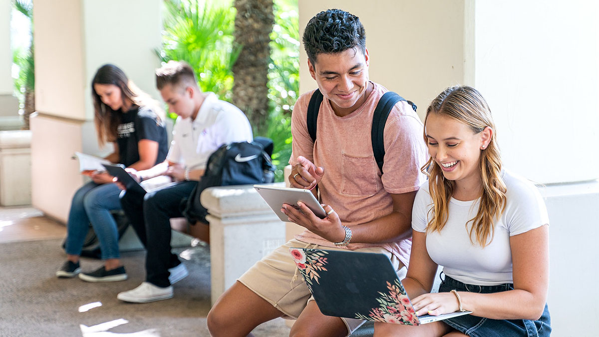 Students sit on benches in a shaded outdoor area, working on laptops and tablets while smiling and studying together.