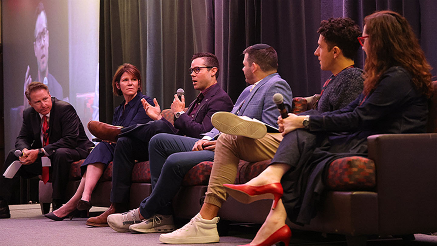 Six panelists seated on stage holding microphones during a live discussion at the AAAI Summit, with one person speaking mid-conversation.